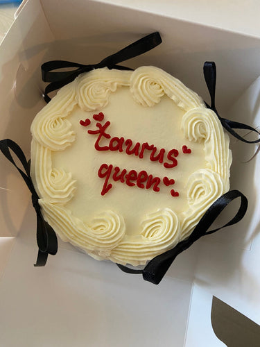 Кръгла бяла торта с кремав кант, панделки и надпис 'taurus queen' на повърхността.
