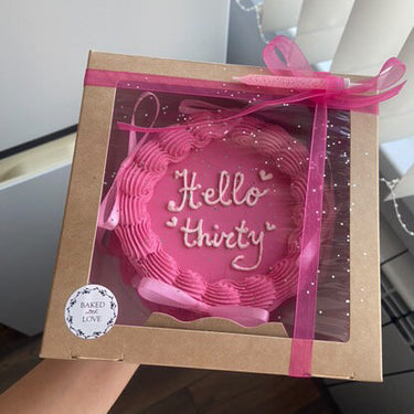 Кръгла розова торта с надпис Hello thirty, декорирана с панделки, кремав кант и брокат.
