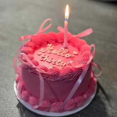 A round pink cake with a cream border, decorative ribbons and the gold inscription "Hello Thirty"