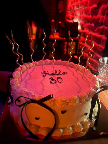 Кръгла бяла торта с надпис 'Hello 30', украсена с панделки и кремав кант по периферията.