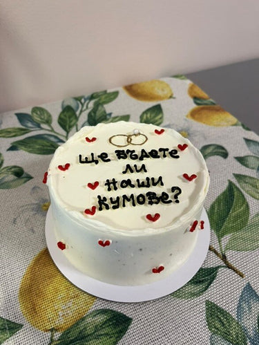 Кръгла бяла торта със златен надпис 'Ще бъдете ли наши кумове?' в елегантен шрифт.
