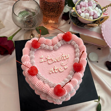 Pink heart-shaped cake with the message 'twenty ate', decorated with cherries and a cream border along the edge.