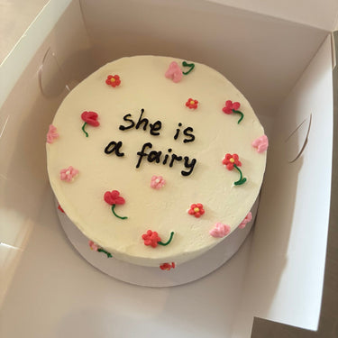 Кръгла бяла торта с рисунка на фея и надпис "she is a fairy" върху глазурата.