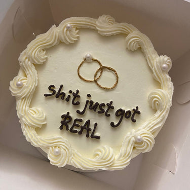 A round cream-coloured wedding cake with pearl decoration, a cream border and the inscription 'shit just got REAL'