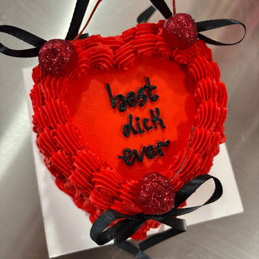 A red heart cake with glitter, ribbons and cherries, inscription 'best dick ever' on top.