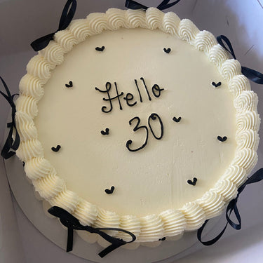Кръгла бяла торта с надпис Hello 30, кремав кант и декоративни панделки.