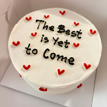 Кръгла бяла торта с надпис 'The Best is Yet to Come' върху глазурата, без допълнителна украса.