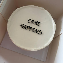 Кръгла бяла торта с надпис CAKE HAPPENS в тъмни букви, без допълнителна украса.