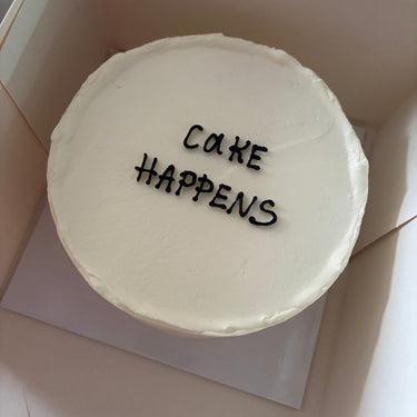 Кръгла бяла торта с надпис CAKE HAPPENS в тъмни букви, без допълнителна украса.