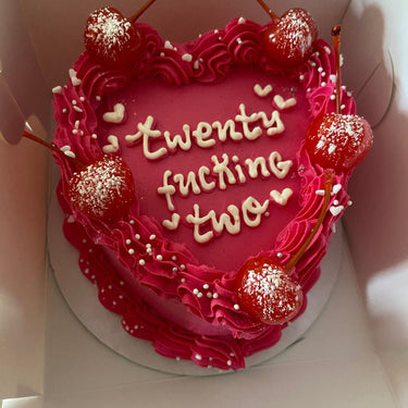Pink heart-shaped cake with cherries, a cream border and pearls. Inscription: twenty fucking two.