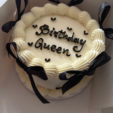 Кръгла бяла торта с надпис Birthday Queen, украсена с панделки и кремав кант.