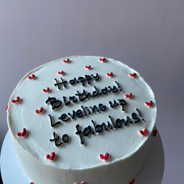 Бяла кръгла торта с надпис 'Happy Birthday! Leveling up to fabulous!' върху глазурата, без допълнителна украса.