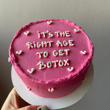 A round pink cake with the inscription 'IT'S THE RIGHT AGE TO GET BOTOX' on top, with no additional decoration.