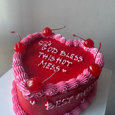 A heart-shaped cake with red icing, cherries, a cream border and the side inscription 'GOD BLESS THIS HOT MESS'.