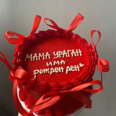 Кръгла червена торта с кремав кант, панделки и надпис 'МАМА УРАГАН има рожден ден' отгоре.
