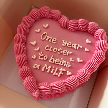 Pink heart-shaped cake with a cream border and the message 'One year closer to being a Milf'