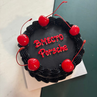 Round cake with black icing, cream border, cherries and the message 'INSTEAD OF a Porsche'