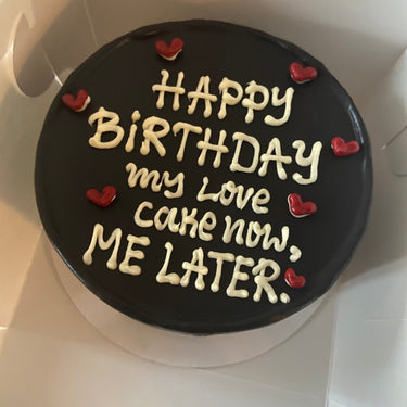 A round black cake with a white inscription 'Happy Birthday my Love cake now, ME LATER'