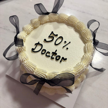 Кръгла бяла торта с панделки, кремав кант и надпис '50% Doctor'.