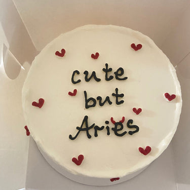 Кръгла бяла торта с надпис 'cute but Aries' на повърхността, без допълнителна украса.